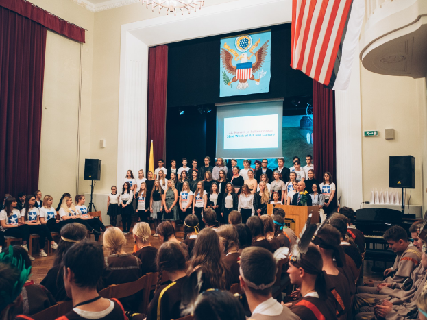 Eine Gruppe von Menschen steht auf einem Podium mit einem Mikrofon, flankiert von Fahnen und Lautsprechern, und spricht eine sitzende Menge bei einem kulturellen Ereignis an.