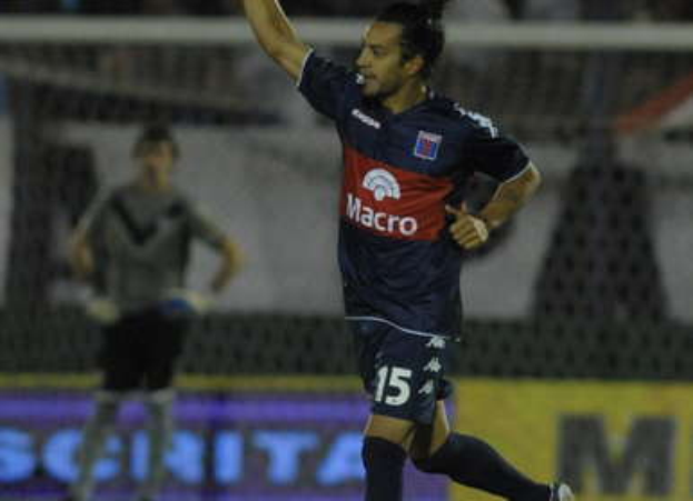 Ein Fußballspieler feiert nach einem Tor auf dem Boden, mit jubelnden Zuschauern und einer Person im Hintergrund, alles vor einem unscharfen, energetischen Hintergrund.