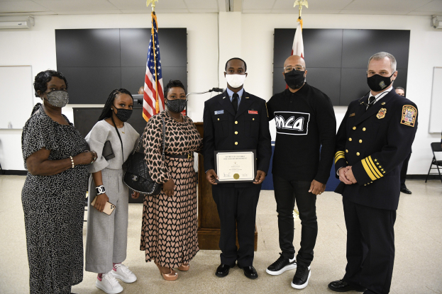 Gruppe von Menschen in Masken vor Flaggen, einer hält ein Zertifikat, während einer Abschlussfeier in einem Raum mit Stühlen, Tischen und Wandtafeln.
