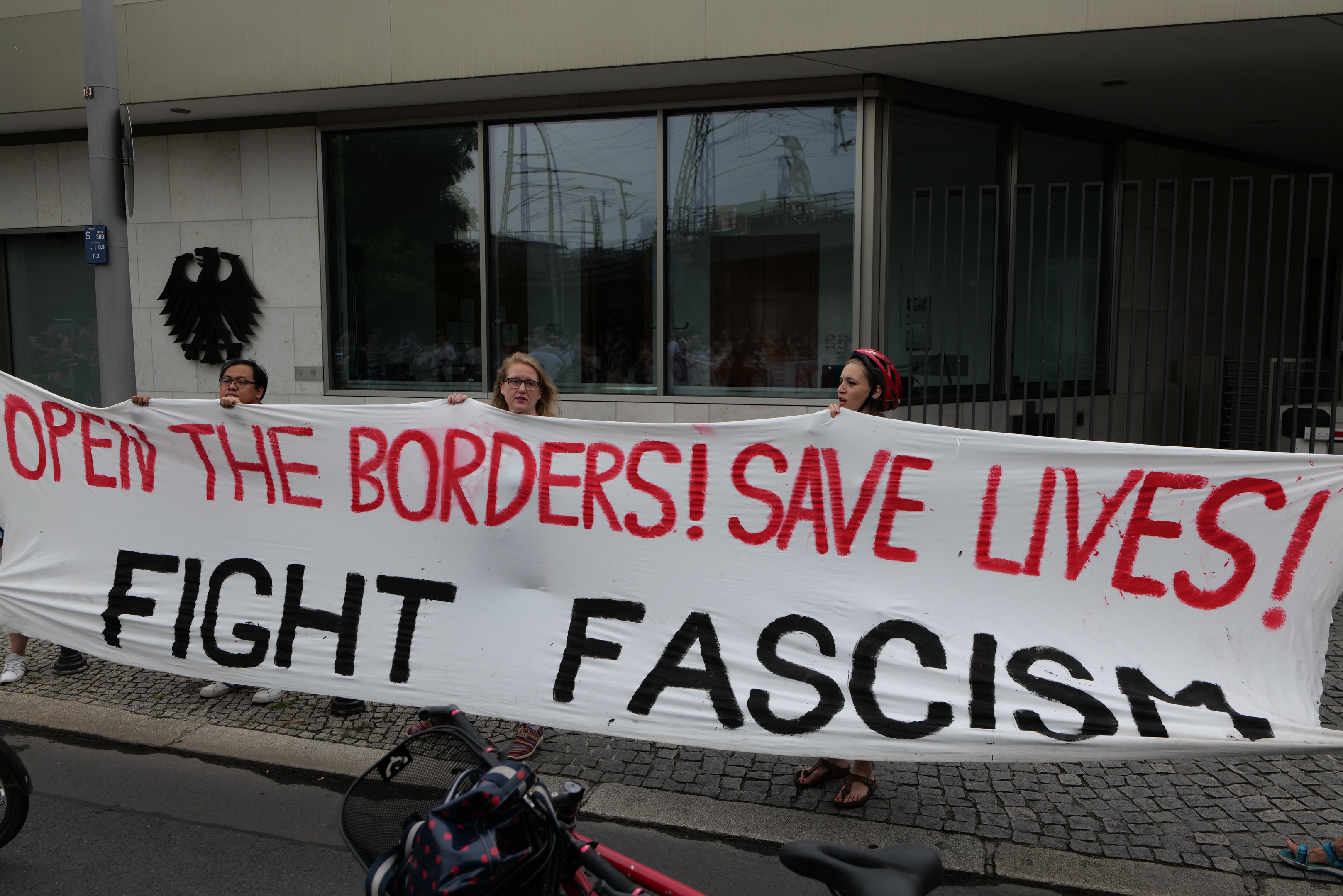 Menschen mit einem Banner mit der Aufschrift "Öffnet die Grenzen, rettet Leben, kämpft gegen Faschismus" vor einem Gebäude mit Glasfenstern und einem Pfahl, mit einem Fahrrad und einer Tasche auf der Straße.