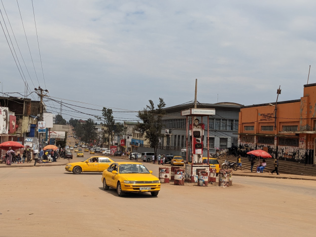 Ein gelbes Taxi fährt auf einer belebten Stadtstraße mit Gebäuden, Passanten mit Schirmen und Strommasten mit Drähten unter einem bewölkten Himmel.