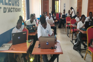 Eine Gruppe von Menschen, die an Tischen mit Laptops in einem Klassenzimmer sitzen, mit Taschen auf dem Boden und Text an der linken Wand, Fenstern und Türen im Hintergrund und Text unten, der auf einen Online-Kurs in Ghana hinweist.