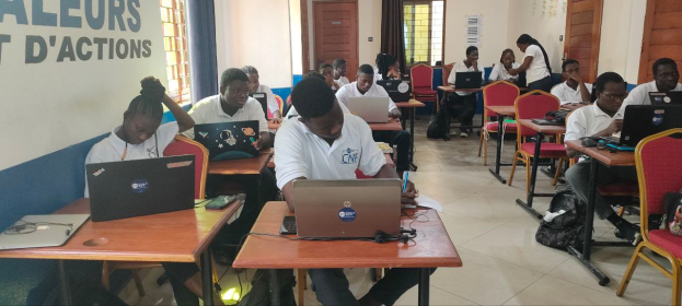 Eine Gruppe von Menschen, die an Tischen mit Laptops in einem Klassenzimmer sitzen, mit Taschen auf dem Boden und Text an der linken Wand, Fenstern und Türen im Hintergrund und Text unten, der auf einen Online-Kurs in Ghana hinweist.