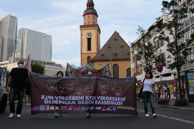 Eine Gruppe maskierter Personen marschiert auf einer Straße und hält ein Transparent, daneben steht ein geparktes Auto, im Hintergrund sind Gebäude, Bäume, Schilder, Pfosten und ein Kirchturm unter einem klaren blauen Himmel zu sehen.