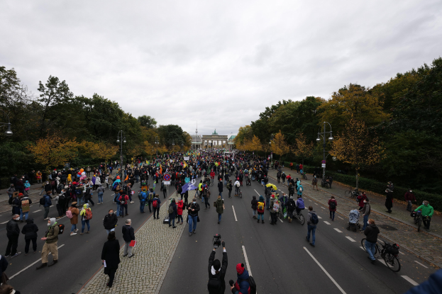 Eine große Gruppe von Menschen marschiert eine baumbestandene Straße mit Laternenpfählen entlang, mit Kameras bewaffnet, mit einem Gebäude und einem klaren Himmel im Hintergrund.