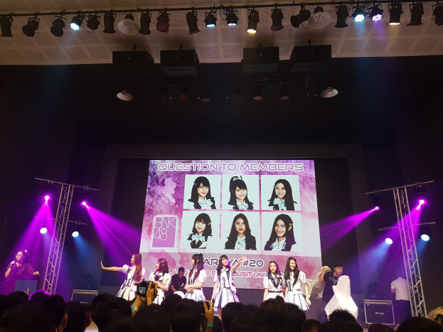 Eine Gruppe von Menschen, die auf einer hell beleuchteten Bühne performen, mit einer Menge darunter, einem Sänger mit Mikrofon und einem Musiker, der ein Instrument spielt, während ein großer Bildschirm Bilder der K-Pop-Gruppe BK48 anzeigt.