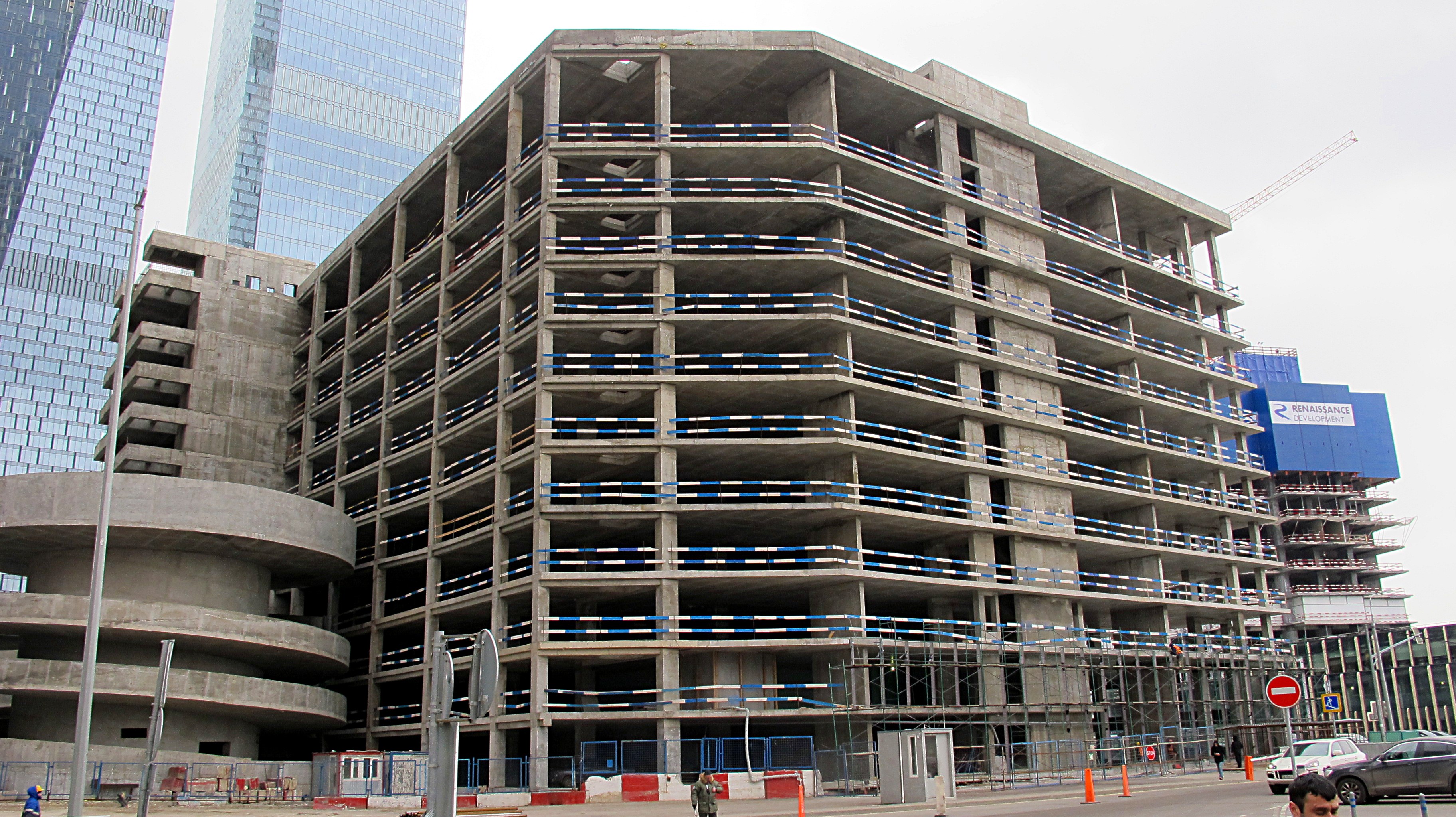 Großes Gebäude im Bau in einer urbanen Gegend mit Wolkenkratzern, Fahrzeugen, Fußgängern, Verkehrs-Infrastruktur und Kränen vor einem klaren Himmel.