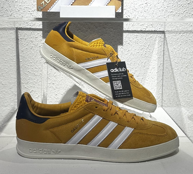 Ein Paar adidas Gazelle OG-Schuhe in Gelb, Weiß und Marineblau auf einer weißen Oberfläche mit einem Anhänger, vor einer weißen Wand mit einem Glasregal im Hintergrund.