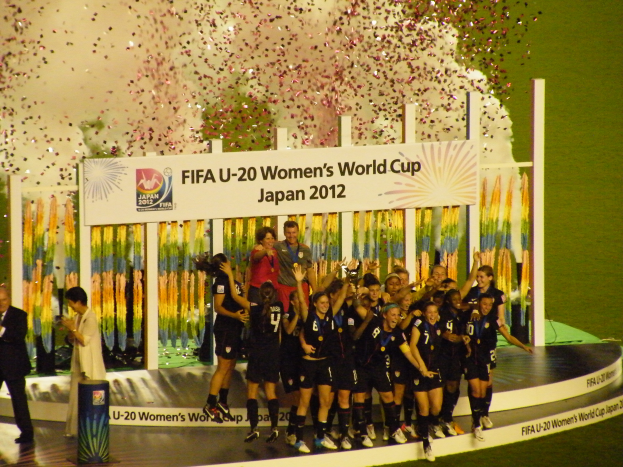 Eine Gruppe von Frauenfußballspielerinnen in schwarzen Uniformen steht auf einer Bühne und feiert ihren Sieg, umgeben von einer Menge mit Kameras, vor einem dekorierten Hintergrund und einem grasbewachsenen Boden.