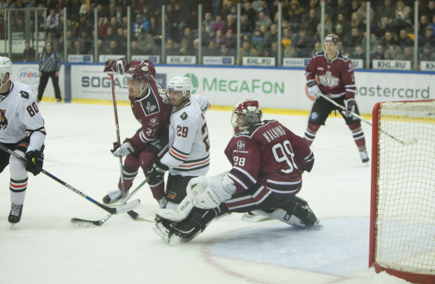 Gruppe von Menschen beim Eishockeyspielen auf einem Eisstadion mit Tor auf der rechten Seite, tragen Helme und halten Stöcke, Zuschauer auf Tribüne mit Bannern im Hintergrund.
