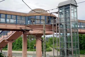 Ein Bahnhof mit einem zentralen Glaselevator, einer umliegenden Brücke mit Säulen und Geländern, Strommasten mit Kabeln, Schildern, einem Zaun, Pflanzen, Bäumen und einer bewölkten Himmel im Hintergrund.