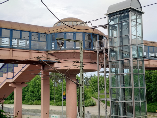 Ein Bahnhof mit einem zentralen Glaselevator, einer umliegenden Brücke mit Säulen und Geländern, Strommasten mit Kabeln, Schildern, einem Zaun, Pflanzen, Bäumen und einer bewölkten Himmel im Hintergrund.