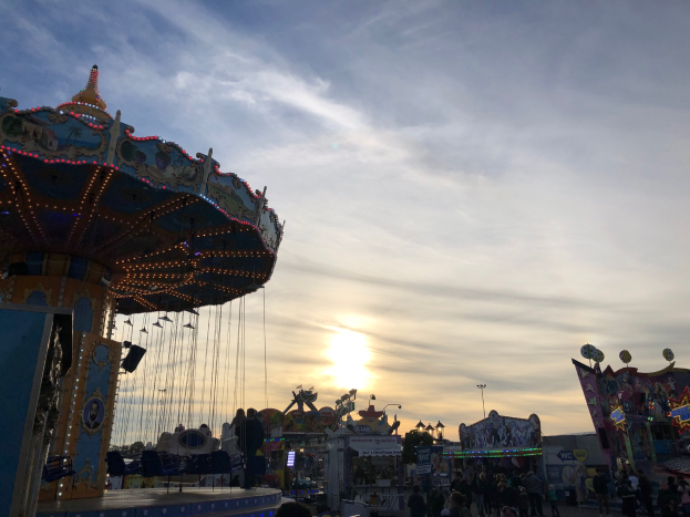 Eine Karussellfahrt auf einem Rummelplatz mit Menschen darauf, umgeben von Rummelplatzattraktionen, Pfählen und Lichtern, unter einem bewölkten Himmel, beleuchtet von der untergehenden Sonne.