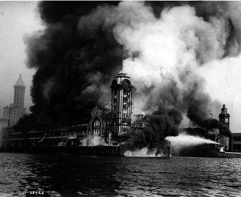 Schwarz-weißes Foto eines großen, von Flammen eingehüllten Gebäudes mit Rauch, der daraus aufsteigt, einem Gewässer im Vordergrund und anderen Gebäuden sowie dem Himmel im Hintergrund.