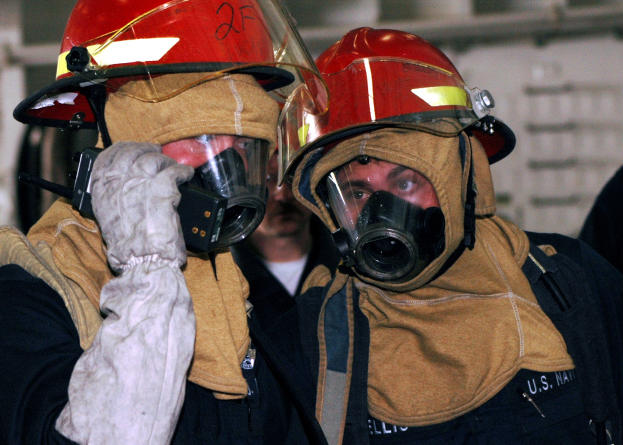 Zwei Feuerwehrleute in Gasmasken und Helmen, einer hält ein Funkgerät in der Hand, stehen neben einer Wand mit Rohren im Hintergrund.