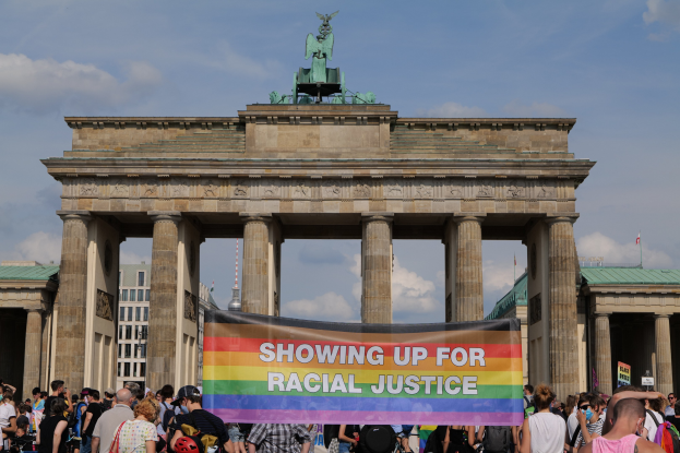 Eine Gruppe von Menschen mit einer "Racial Justice"-Schriftzug-Tafel vor dem Brandenburger Tor in Berlin, mit den Säulen und der Statue des Tors.