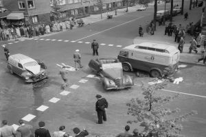 Schwarz-weiß-Szene eines Autounfalls an einer Straßenkreuzung mit einem Lieferwagen, anderen Fahrzeugen und einer Gruppe von Menschen in der Nähe, umgeben von Gebäuden, Bäumen, Pfählen und Texttafeln.