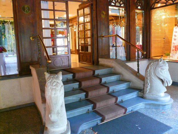 Zwei steinerne Pferdestatuen flankieren den Teppichboden des Eingangs eines Geschäfts mit Glastüren, Schaufensterpuppen in Kleidern und Innenbeleuchtung.