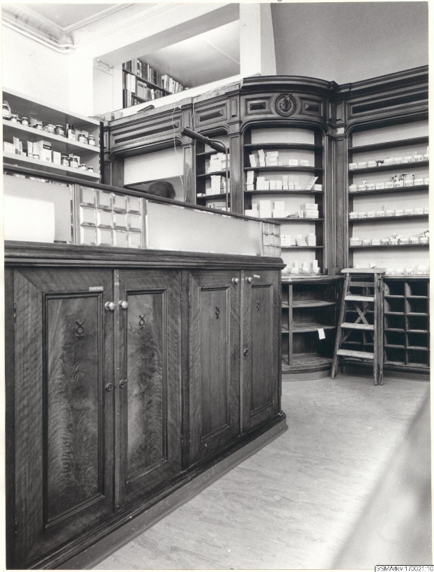 Schwarzes und weißes Foto eines vintage Apothekeninneren mit einer Theke, Schränken, einer Leiter, Regalen voller Behälter und undeutlicher Schrift oben.