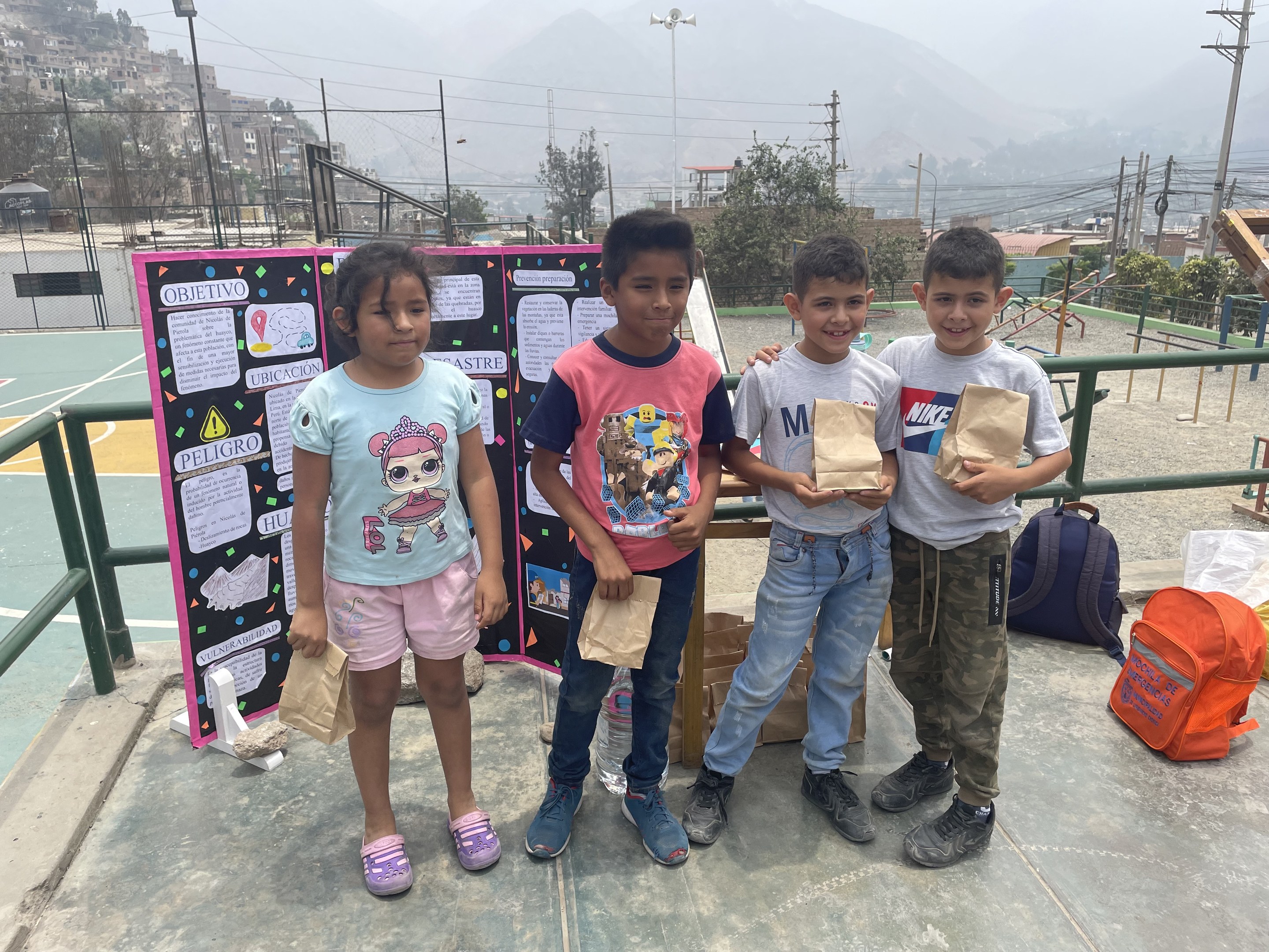 Gruppe von Kindern auf einem Basketballfeld mit Papierbeuteln in den Händen, mit Beuteln und Gegenständen auf der rechten Seite und einer Tafel mit Text auf der linken Seite, vor einem Hintergrund aus Zäunen, Pfosten, Straßenlaternen, Bäumen, Gebäuden, Höhen und Himmel.