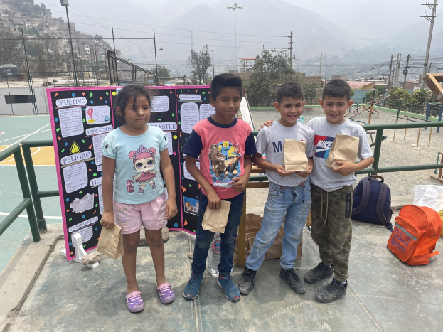 Gruppe von Kindern auf einem Basketballfeld mit Papierbeuteln in den Händen, mit Beuteln und Gegenständen auf der rechten Seite und einer Tafel mit Text auf der linken Seite, vor einem Hintergrund aus Zäunen, Pfosten, Straßenlaternen, Bäumen, Gebäuden, Höhen und Himmel.