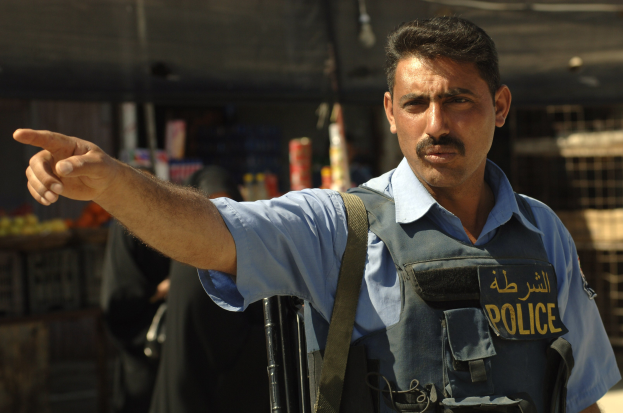 Polizeibeamter in Uniform mit Pistole vor einem Geschäft stehend, mit unscharfen Menschen und Gegenständen im Hintergrund.