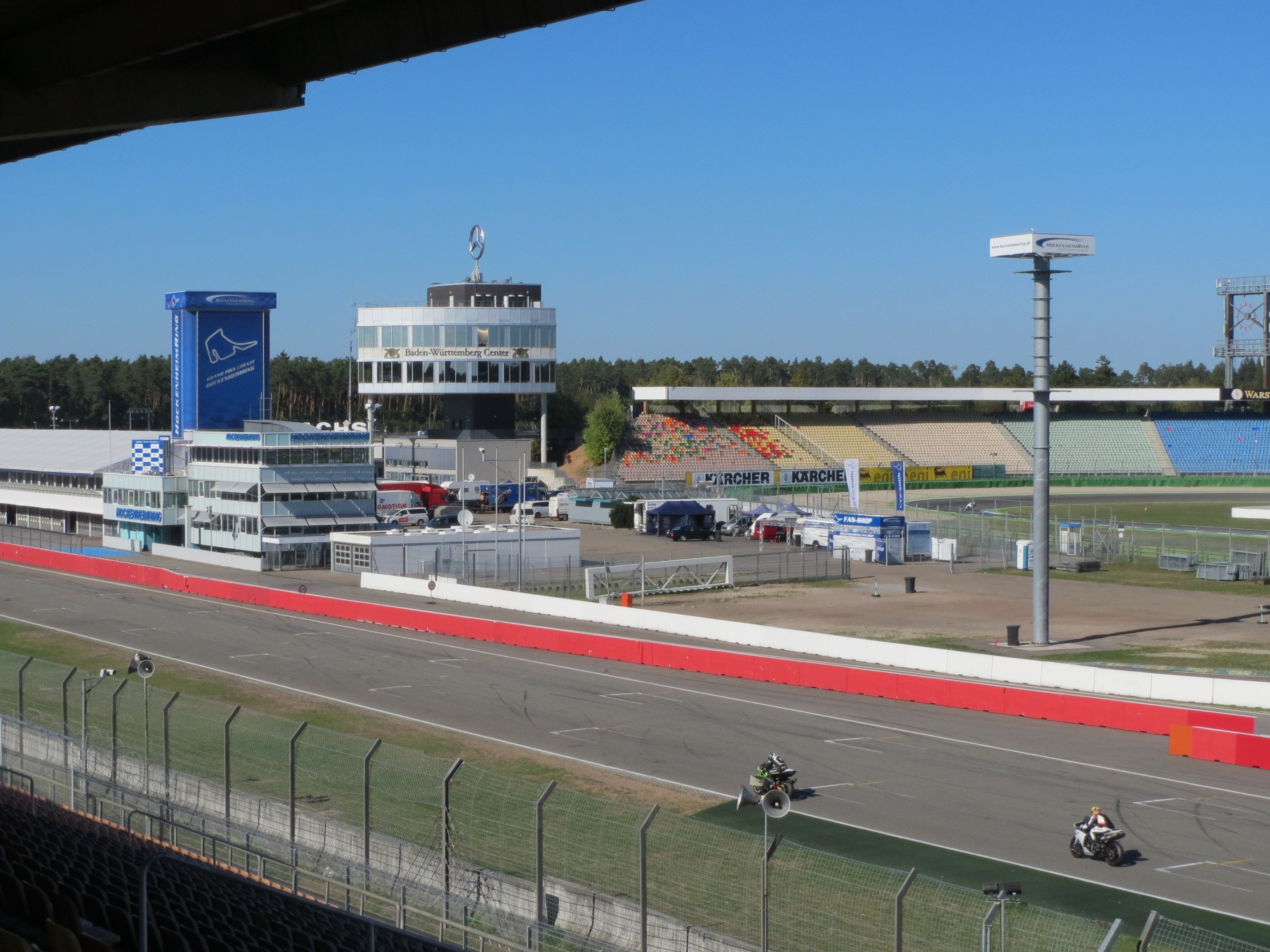 Gruppe von Motorradfahrern auf einer Rennstrecke, umgeben von einem Metallzaun, Gras, Pfählen und einem Stadion unter einem bewölkten Himmel.