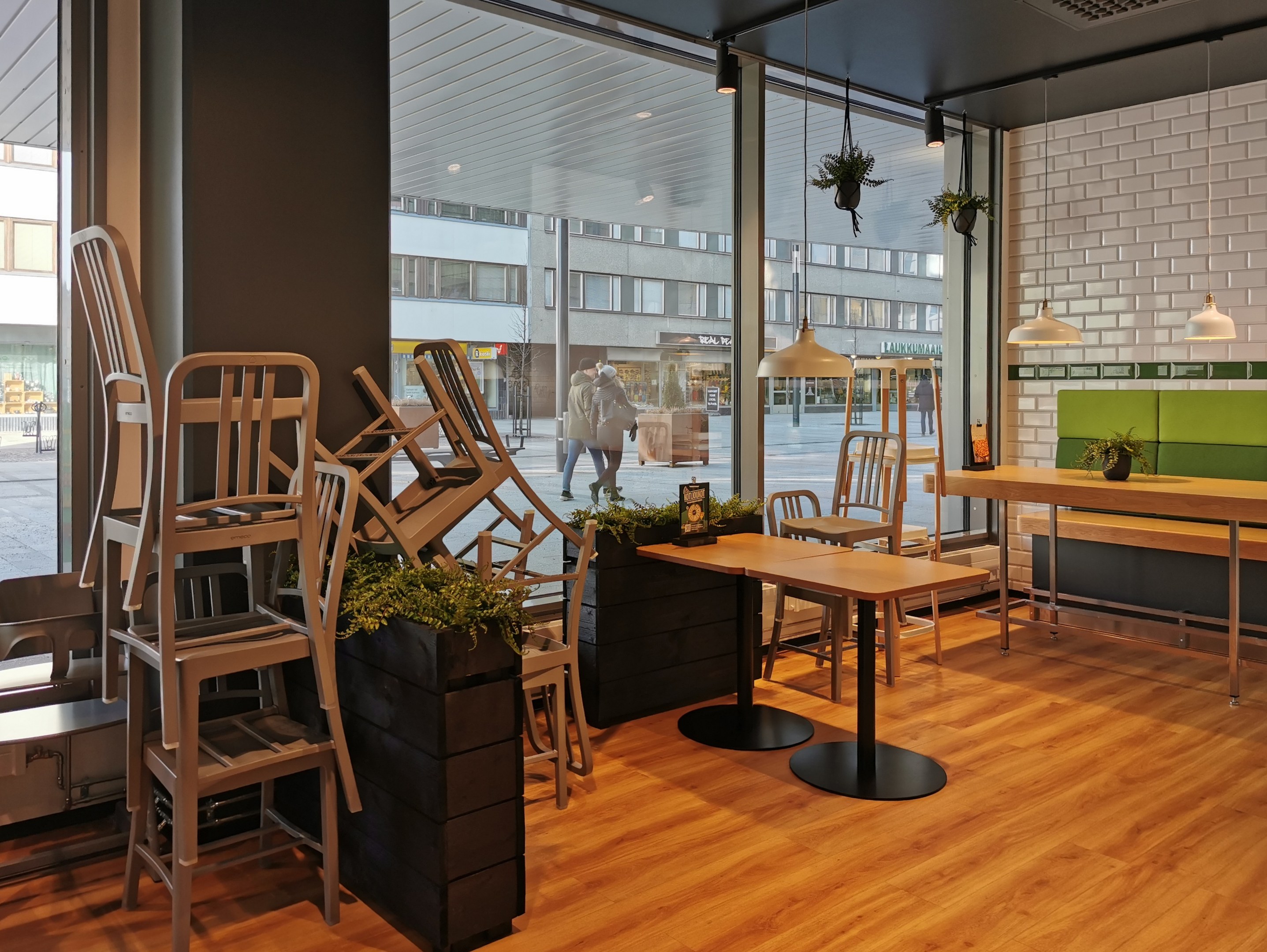 Ein Restaurant-Innenraum mit Holztischen und -stühlen in der Nähe eines großen Fensters, das auf eine Berliner Straße mit Passanten, Gebäuden und urbanen Elementen blickt.