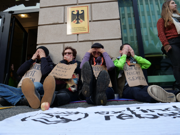 Menschen sitzen vor einem Gebäude auf dem Boden und halten Schilder, mit einer Fahne auf der Straße und einer Frau stehend rechts daneben, bei einer scheinbaren Protestaktion gegen eine Regierungsentscheidung.