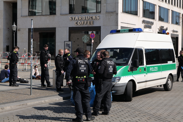 Polizeibeamte vor einem Starbucks-Café mit einem Van auf der rechten Seite und Menschen auf der linken Seite sowie einem Gebäude, einem Schild, einem Laternenmast und einem Zaun im Hintergrund.