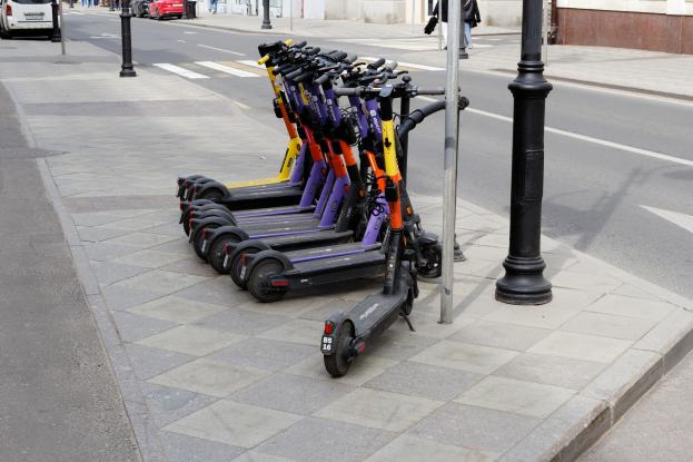 Eine Reihe von Elektro-Scootern, die entlang einer Straße geparkt sind, mit fahrenden Fahrzeugen, Passanten auf dem Gehweg und Gebäuden im Hintergrund.