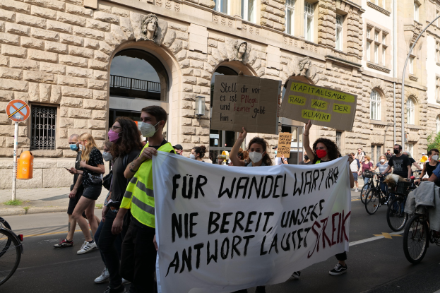 Eine Gruppe von Menschen marschiert bei einer Demonstration eine Straße entlang, einige halten Schilder und andere fahren Fahrräder, mit einem Gebäude im Hintergrund, das Fenster, Bögen, Säulen und Skulpturen aufweist.