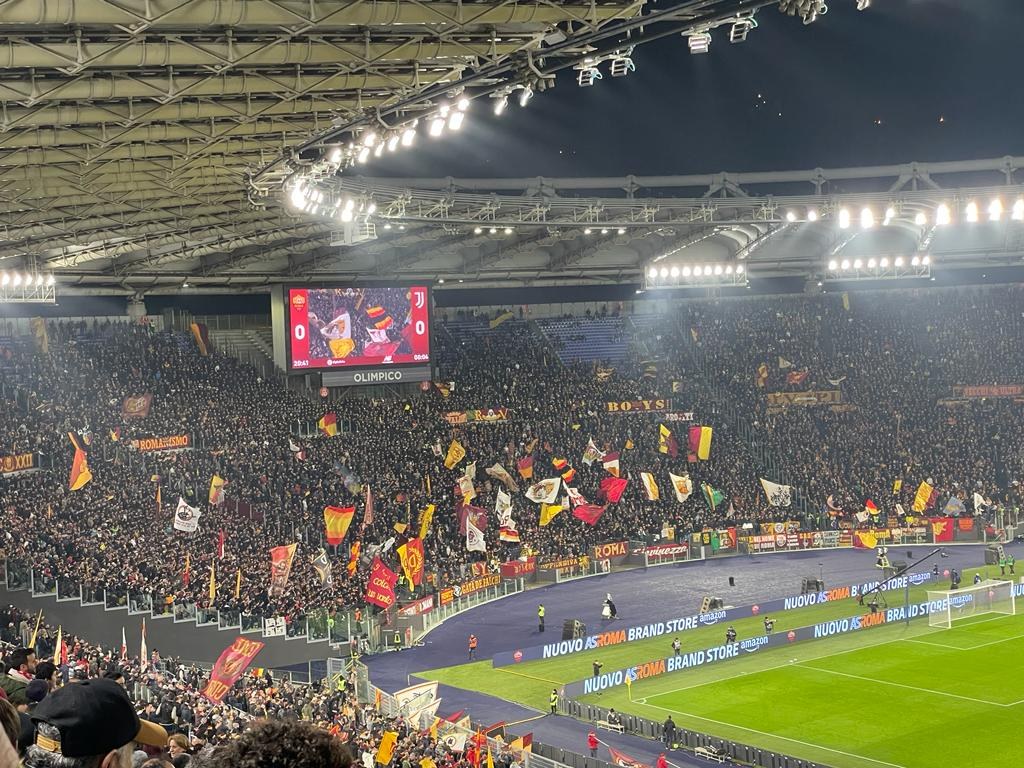 Eine große Menge Menschen in einem Stadion schwenkt Fahnen und Banner, mit Menschen in Mützen auf den Tribünen unter beleuchteten Dächern, die auf ein Fußballspiel warten.