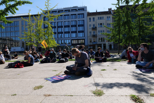 Eine Gruppe von Menschen sitzt vor einem Gebäude in Berlin auf dem Boden, einige tragen Masken, mit verstreuten Taschen und Gegenständen, unter einem klaren blauen Himmel umgeben von Bäumen.