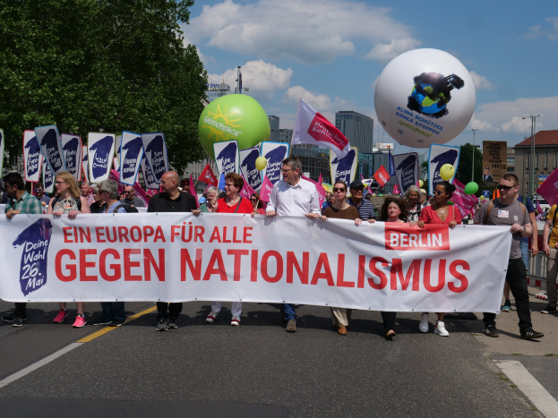 Eine Gruppe von Menschen marschiert bei einer Demonstration gegen Nationalismus in Berlin, hält ein Banner, Fahnen und Ballons, mit Bäumen, Gebäuden und einem bewölkten Himmel im Hintergrund.