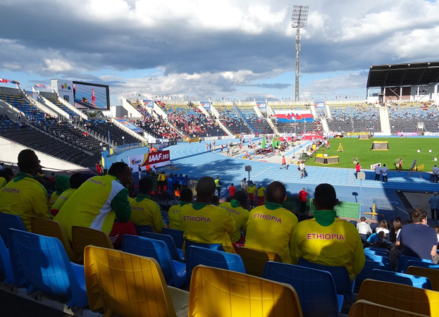 Schwarzes und weißes Foto einer vollen Tribüne mit Zuschauern bei einem Fußballspiel, mit Bannern, Pfosten, einem Schuppen, Bäumen, einem Turm und einem bewölkten Himmel.