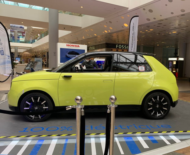 Gelber Wasserstoff-elektrischer Auto vor einem Gebäude geparkt, umgeben von Barrieren und Bannern, mit Menschen in der Nähe und Deckenventilatoren oben.