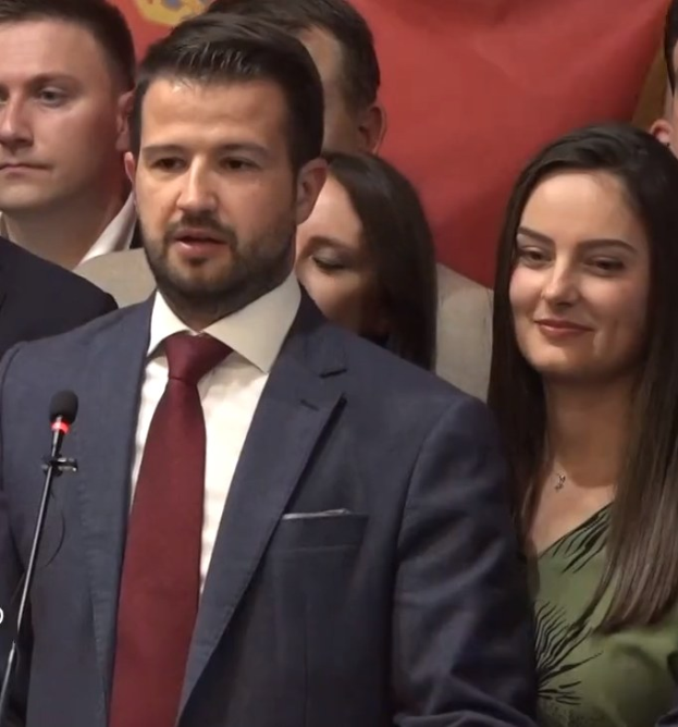Ein Mann im Anzug spricht in ein Mikrofon auf einem Podium, mit einem Banner dahinter und einer Menge davor, wahrscheinlich zu einem Regierungsverbot von sozialen Medien.