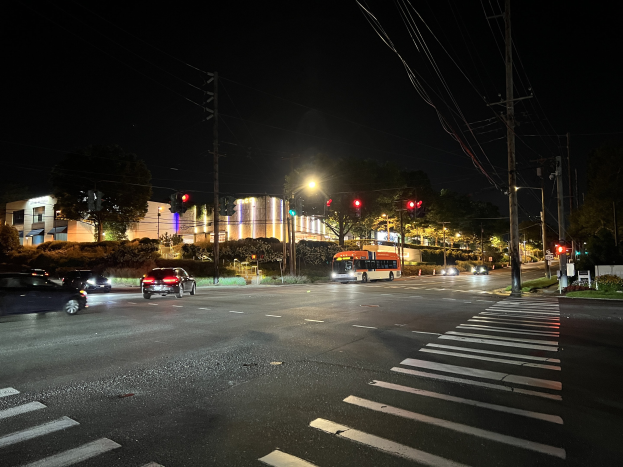 Eine nächtliche Stadtstraße mit fahrenden Autos und einem Bus, beleuchtet von Straßen- und Ampellicht, umgeben von Gebäuden, Bäumen und Gras unter einem dunklen Himmel.
