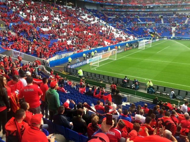 Eine große Menschenmenge in einem Stadion schaut einem Fußballspiel zu, mit Sitzplätzen und Stehplätzen, Bannern, einem Torpfosten und einer Decke mit Leuchten.