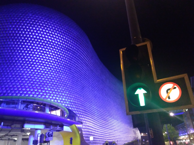 Das Selfridges Einkaufszentrum in Birmingham, England, ein Gebäude mit Säulen und Lichtern, umgeben von Bäumen, Laternenmasten und Fahrzeugen auf der Straße, mit einer Ampel davor und dem Himmel im Hintergrund.