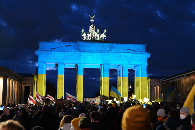 Menschenmenge mit Fahnen und Plakaten vor dem Brandenburger Tor, mit einem Banner auf der rechten Seite und bewölktem Himmel darüber.