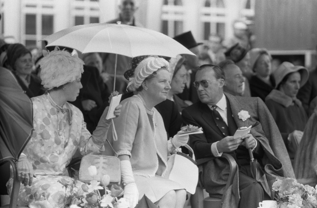 Schwarzes und weißes Foto von Queen Elizabeth II und Prinz Philip, die mit Schirmen sitzen, umgeben von Menschen mit Hüten und Büchern, mit einem Gebäude und Blumenschmuck im Hintergrund.