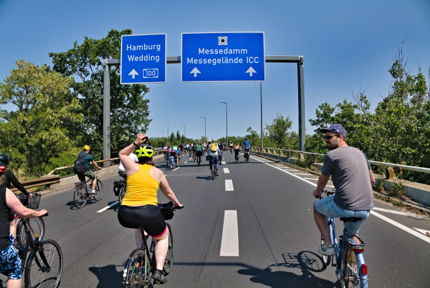 Gruppe von Radfahrern in Helmen auf einer Straße mit einem Geländer und Bäumen auf beiden Seiten, Laternen im Hintergrund, unter einem klaren blauen Himmel, mit einem Schild, das eine Radtour in Hamburg anzeigt.