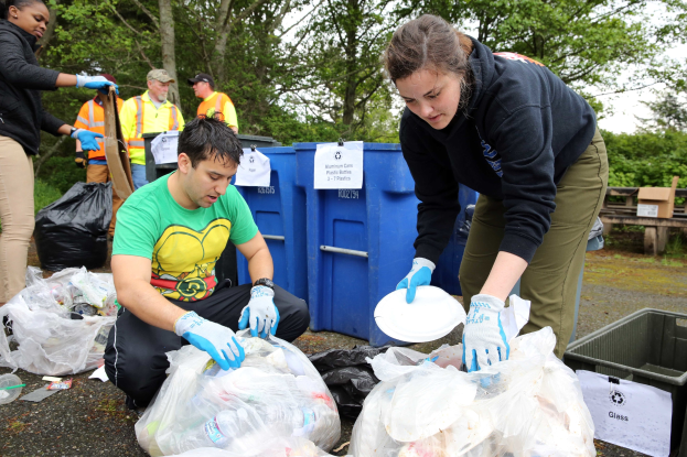 Eine Gruppe von Menschen im Park Müll sammeln, darunter ein Mann und eine Frau mit Handschuhen, die Teller halten, umgeben von Müll wie Plastikabdeckungen und Flaschen, mit einem Müllcontainer und einer Bank rechts und Bäumen und einem blauen Himmel im Hintergrund.