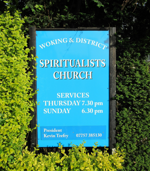 Ein Schiefertafel-Schild mit der Aufschrift "Woking & District Spiritualists Church" wird draußen zwischen Grünpflanzen und einem Gebäude aufgestellt.