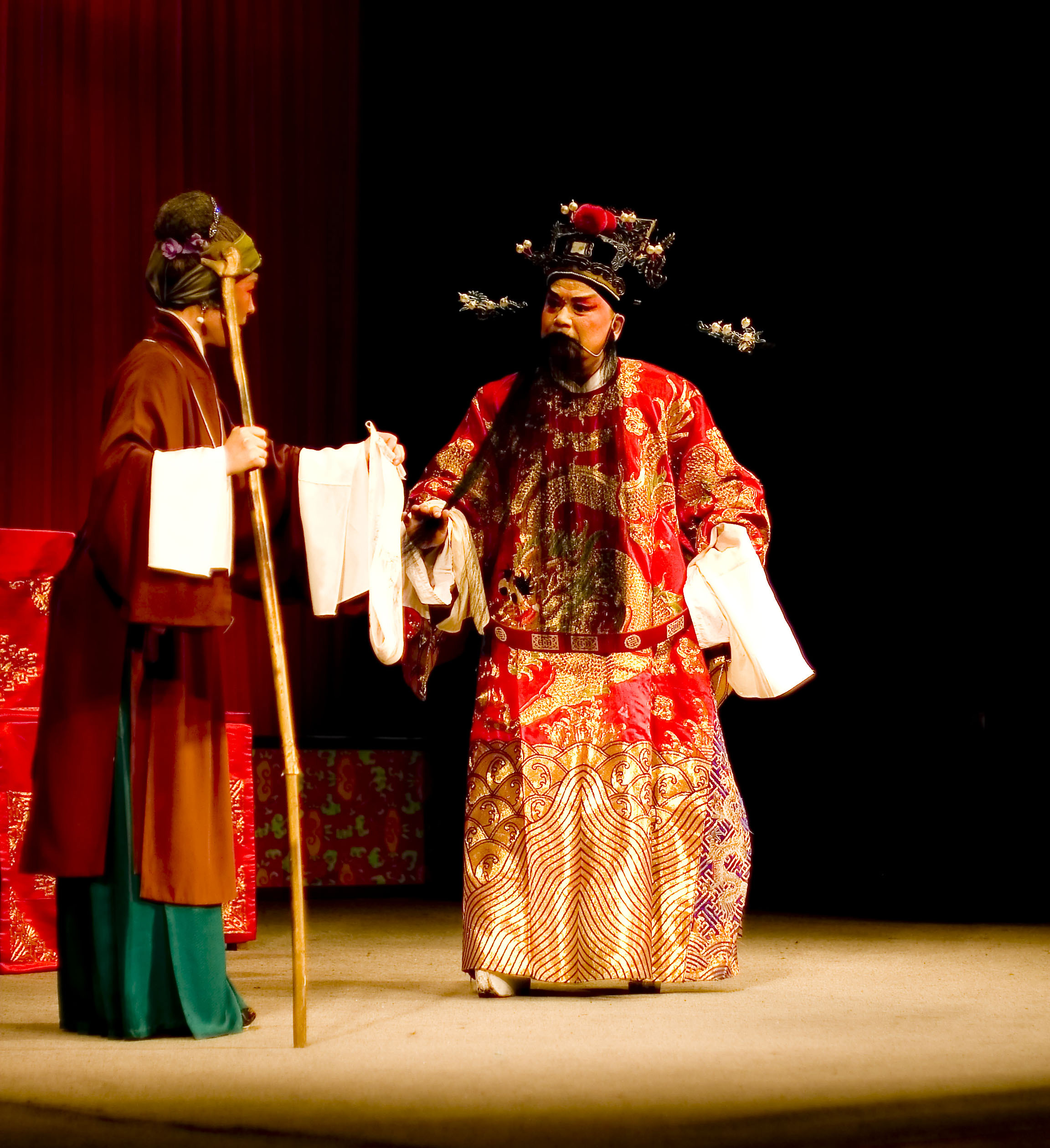 Ein Mann in einem roten Gewand und eine Frau in einem grünen Kleid stehen auf einer Bühne mit einem roten Vorhang dahinter; der Mann hält einen Stock und Papiere.