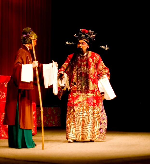 Ein Mann in einem roten Gewand und eine Frau in einem grünen Kleid stehen auf einer Bühne mit einem roten Vorhang dahinter; der Mann hält einen Stock und Papiere.