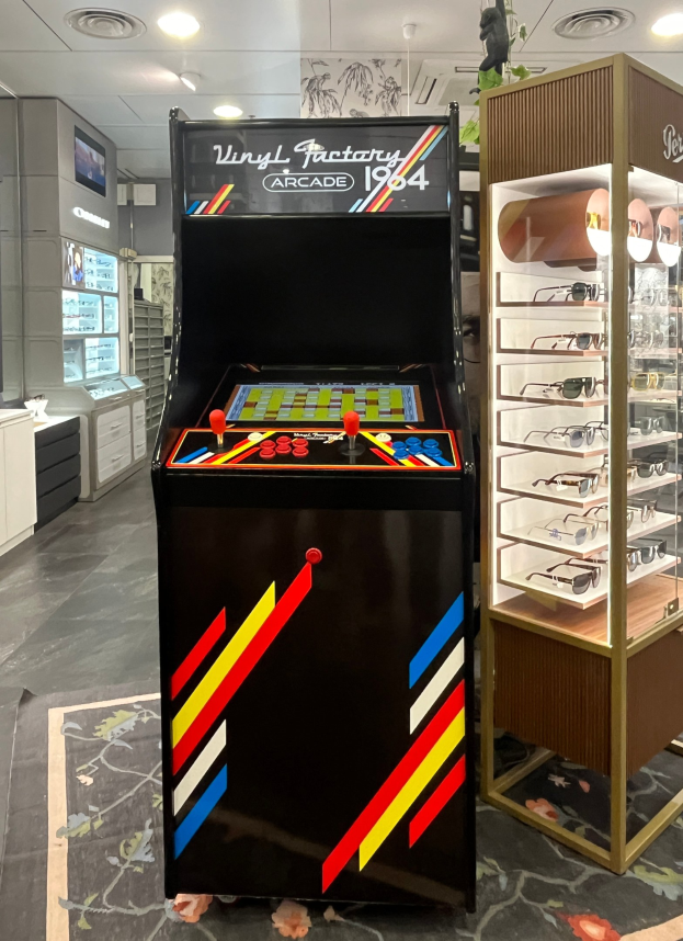 Ein Geschäft mit einem alten Arcade-Automaten in der Mitte, einer Glasvitrine auf der rechten Seite und Retro-Beleuchtung mit Schränken und einem Fernseher im Hintergrund.