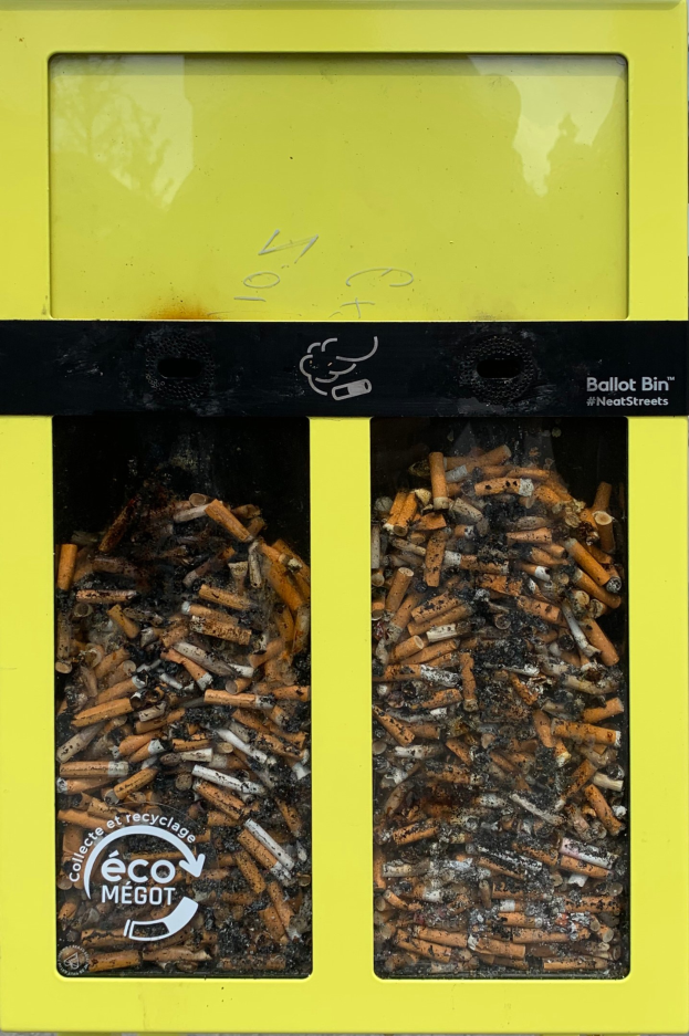 Ein leuchtend gelber Recyclingmüllcontainer voller Zigarettenstummel auf einem Gehweg, beschriftet für die Entsorgung von Zigarettenabfall.